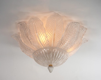 May include: A flush mount ceiling light fixture featuring clear glass panels shaped like large, textured leaves arranged in a circular pattern. The center base is a fluted glass bowl with a small, pointed finial at the bottom, emitting a warm glow.
