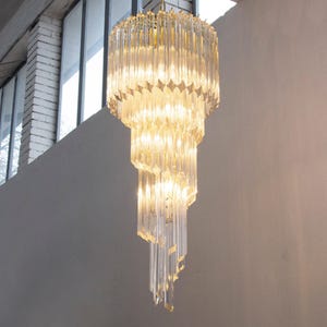 May include: A multi-tiered chandelier featuring numerous vertical, clear glass prisms arranged in a cascading spiral formation. The structure is composed of several circular levels that decrease in diameter toward the bottom, creating a tapered, elongated silhouette that reflects light through the transparent glass elements.