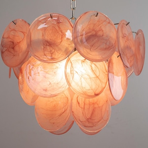 May include: A chandelier featuring multiple circular, translucent peach-colored glass discs with a swirling, marbled pattern. The discs are arranged in a layered, spherical cluster around a central light source, suspended by a silver-toned metal chain.