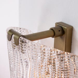 a wall-mounted light fixture with a clear, textured glass shade. The fixture has a brass-colored metal bracket and is attached to a white wall.