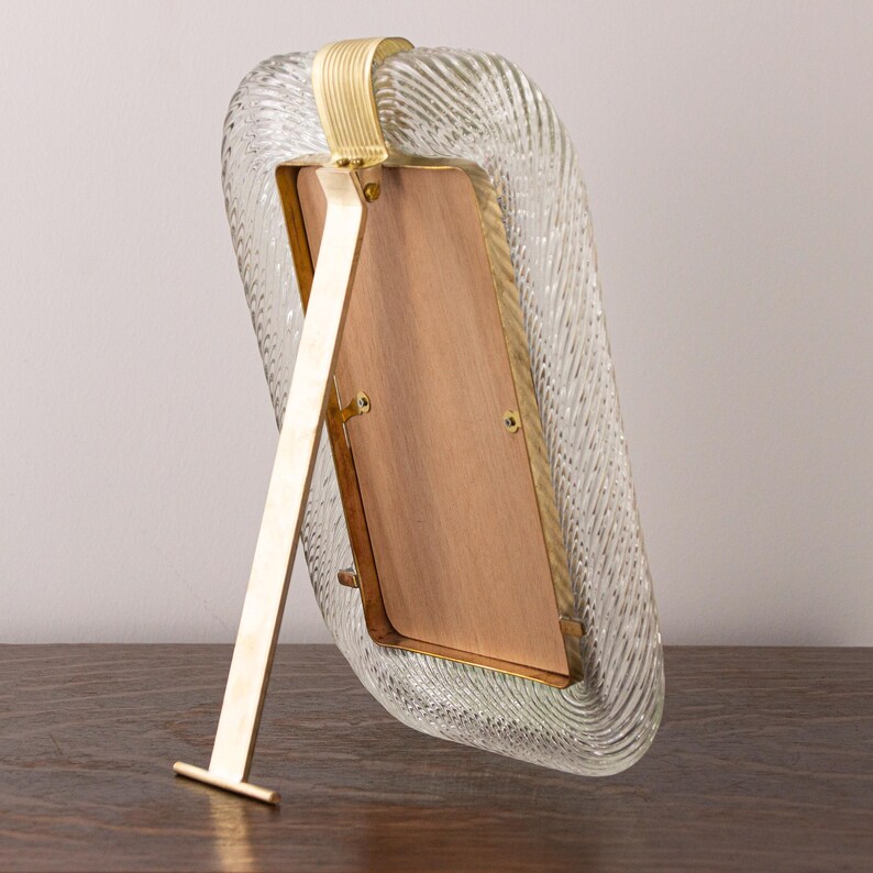May include: A gold-toned picture frame with a clear, textured glass front. The frame has a kickstand on the back to stand upright.