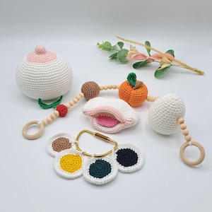 Puede incluir: Un conjunto de juguetes para bebés de crochet con una variedad de texturas y colores. El conjunto incluye una bola blanca de crochet con un centro rosa, una naranja de crochet con un tallo verde, una concha de crochet con un interior rosa, un huevo blanco de crochet y un conjunto de cinco círculos de crochet en diferentes colores.