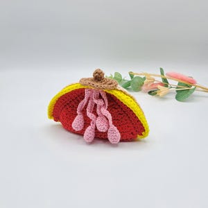 Puede incluir: Un bolso de crochet con forma de semicírculo con un exterior rojo y amarillo y detalles de crochet rosa. El bolso tiene una parte superior de crochet marrón.
