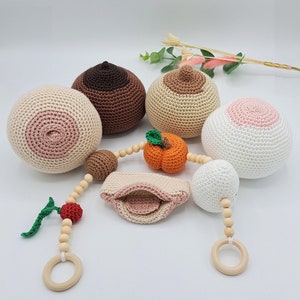 Puede incluir: Una colección de artículos de ganchillo hechos a mano, que incluyen varias esferas con forma de pecho en varios tonos de piel, una calabaza y una pieza con forma de boca. Los artículos son en tonos beige, marrón y blanco, con acentos rosas. También hay una guirnalda de cuentas de madera con una cereza y un anillo de madera.