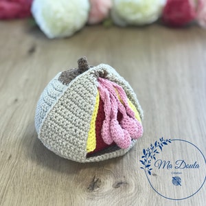 Puede incluir: Un juguete de peluche de ganchillo multicolor con forma de capullo de flor. El juguete está hecho con hilo beige, rosa, amarillo y rojo. El juguete está sobre una superficie de madera.