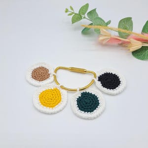 Puede incluir: Un llavero dorado con cuatro círculos de ganchillo unidos. Los círculos son blancos con un centro marrón, amarillo, verde y negro.