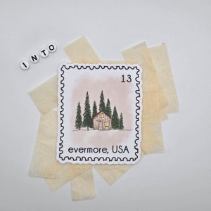 May include: A white sticker with a black border and a drawing of a cabin in the woods. The sticker says "evermore, USA" and "13" in the top right corner.