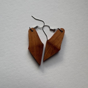 May include: A pair of wooden earrings with a geometric design. The earrings are a warm brown color and have silver-colored ear wires. The wooden pieces are shaped like abstract triangles.