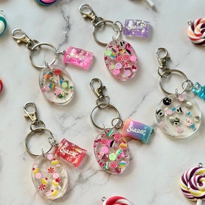 May include: A collection of clear resin keychains with candy-themed charms. Each keychain features a silver clasp and ring, with colorful candy pieces and miniature candy wrappers. Various lollipop charms are scattered around the keychains.