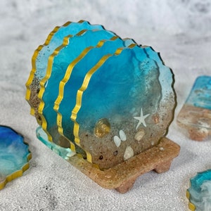 May include: Set of four blue and gold resin coasters with a beach theme. The coasters feature a sandy bottom with seashells and a starfish. The coasters are stacked on a brown resin stand.