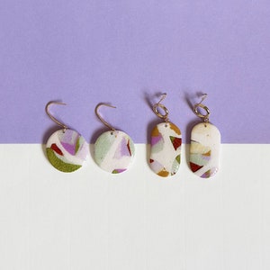 Peut inclure: Quatre paires de boucles d'oreilles avec des motifs abstraits dans des tons de blanc, vert, violet et marron. Les boucles d'oreilles sont en céramique et ont des crochets en métal doré.