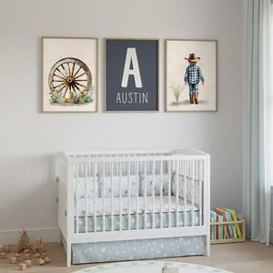 Può includere: Una scena di asilo nido con una culla bianca con biancheria da letto blu e bianca a forma di stella e una gonna per culla abbinata. Sopra la culla ci sono tre stampe incorniciate: una ruota di carro, il nome "Austin" con una grande lettera "A" e un bambino in abiti da cowboy.
