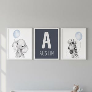 May include: Three framed nursery art prints. One features an elephant holding a blue balloon, another a zebra with a blue balloon, and the centre print has the letter "A" and the name "Austin". The prints are in white frames.