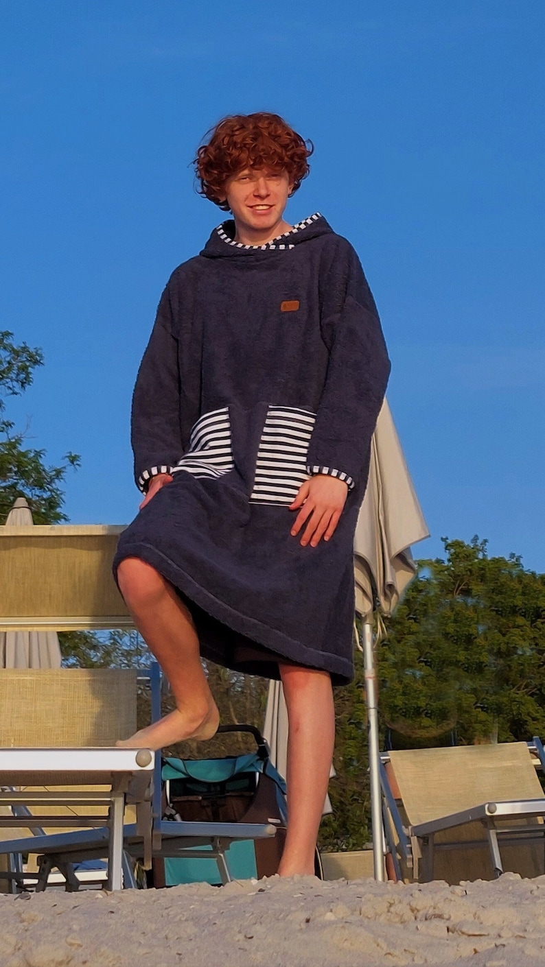 May include: A person wearing a dark blue hooded towel with a white and black striped pocket. The towel has a brown patch on the front. The person is standing on a sandy beach with beach chairs and a stroller in the background.