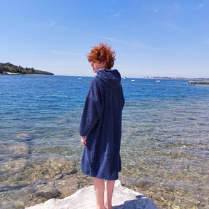 May include: A person wearing a blue hooded towel stands on a rock by the water's edge. The water is clear and shallow, with a small island in the distance.