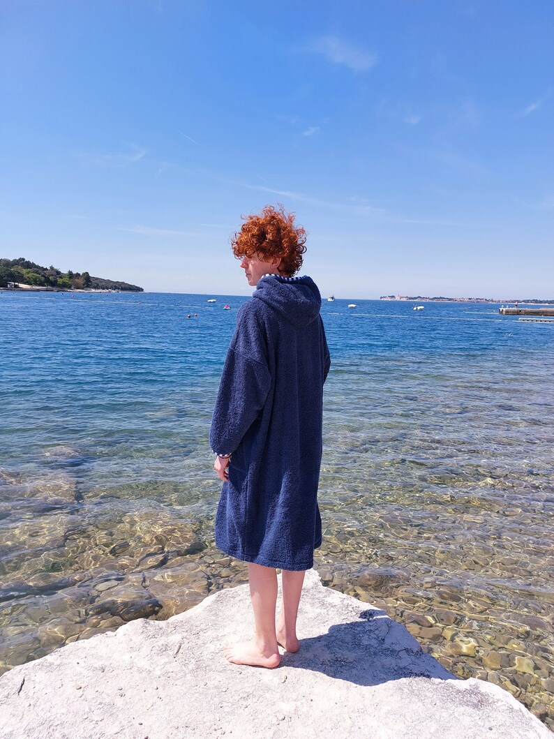 May include: A person wearing a blue hooded towel stands on a rock by the water's edge. The water is clear and shallow, with a small island in the distance.