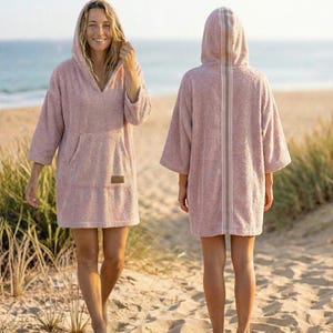Könnte beinhalten: Zwei rosa Strandponchos mit Kapuze. Die Ponchos haben eine Vordertasche und eine Kapuze. Der Poncho auf der rechten Seite hat einen vertikalen Streifen auf der Rückseite. Die Ponchos sind aus weichem, saugfähigem Material. Der Hintergrund ist ein Sandstrand und das Meer.