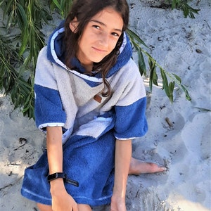 May include: A young person wearing a blue and white hooded towel, sitting on a sandy beach. The towel has a white hood with a blue trim and a blue body with a white pattern. The person is wearing a black fitness tracker on their wrist.