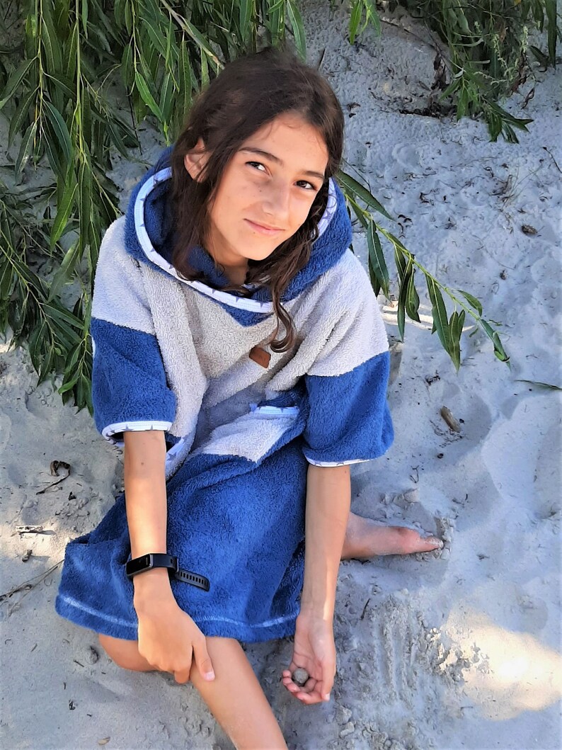 May include: A young person wearing a blue and white hooded towel, sitting on a sandy beach. The towel has a white hood with a blue trim and a blue body with a white pattern. The person is wearing a black fitness tracker on their wrist.