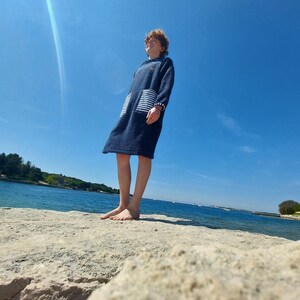 May include: A young person wearing a blue hooded towel with white stripes stands on a rocky shore, looking up at the bright blue sky.