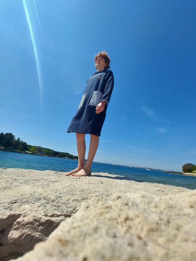 May include: A young person wearing a blue hooded towel with white stripes stands on a rocky shore, looking up at the bright blue sky.