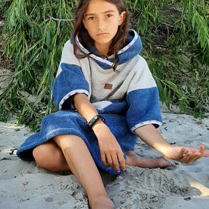 May include: A young person wearing a blue and white hooded towel sits on a sandy beach. The towel has a white stripe around the hood and sleeves.