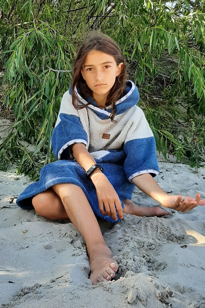 May include: A young person wearing a blue and white hooded towel sits on a sandy beach. The towel has a white stripe around the hood and sleeves.