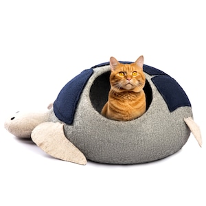 Puede incluir: Un gato jengibre se sienta dentro de una cama para mascotas con forma de tortuga gris y azul. La cama tiene una textura suave y mullida y una abertura redonda para que el gato pueda entrar.