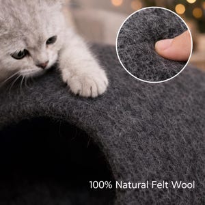 Peut inclure: Une grotte pour chat en laine feutr&eacute;e 100 % naturelle de couleur gris fonc&eacute; avec une entr&eacute;e en tunnel arrondie. Un chaton clair pose sa patte sur le dessus de la structure, tandis qu'un encart circulaire montre la texture fibreuse de la laine.