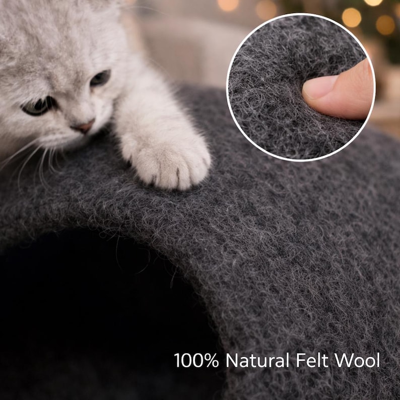 Peut inclure: Une grotte pour chat en laine feutr&eacute;e 100 % naturelle de couleur gris fonc&eacute; avec une entr&eacute;e en tunnel arrondie. Un chaton clair pose sa patte sur le dessus de la structure, tandis qu'un encart circulaire montre la texture fibreuse de la laine.