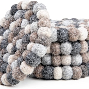 May include: A stack of felt ball coasters in shades of gray, brown, and cream. The coasters are made of small, tightly packed felt balls, creating a textured surface. The design is circular, with a slightly curved shape.