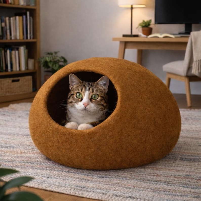 Peut inclure: Une grotte pour animal de compagnie en feutre marron, de forme ronde avec une grande ouverture circulaire. Un chat tigr&eacute; aux yeux verts et aux marques blanches sur le visage et le poitrail est assis &agrave; l'int&eacute;rieur, regardant vers l'avant.