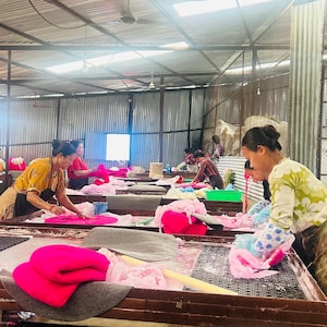 Peut inclure: Un atelier int&eacute;rieur avec plusieurs longues tables o&ugrave; des ouvriers traitent des mat&eacute;riaux en laine feutr&eacute;e rose vif et gris. L'espace de travail poss&egrave;de un toit en m&eacute;tal ondul&eacute; et des c&ocirc;t&eacute;s ouverts, avec divers tas de fibres textiles color&eacute;es.