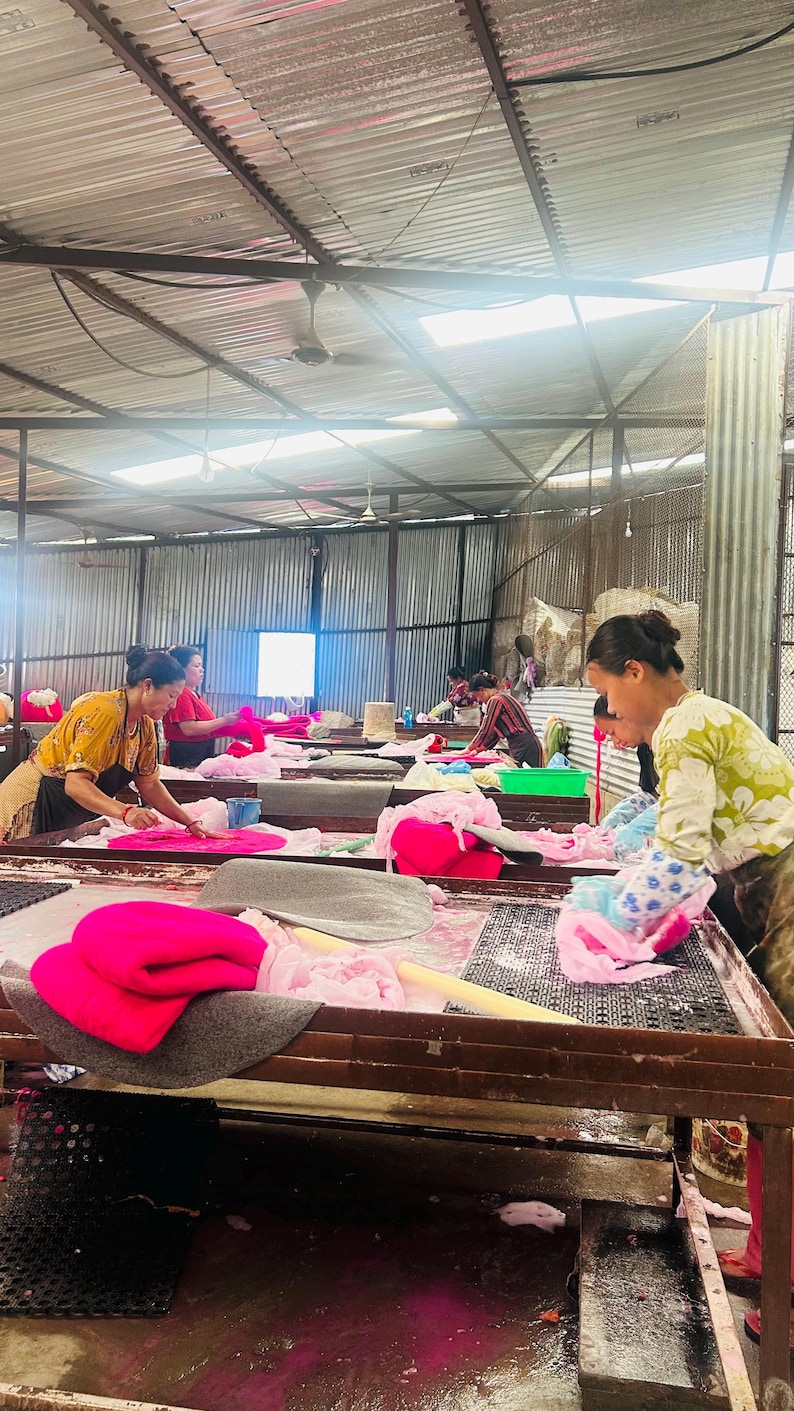 Peut inclure: Un atelier int&eacute;rieur avec plusieurs longues tables o&ugrave; des ouvriers traitent des mat&eacute;riaux en laine feutr&eacute;e rose vif et gris. L'espace de travail poss&egrave;de un toit en m&eacute;tal ondul&eacute; et des c&ocirc;t&eacute;s ouverts, avec divers tas de fibres textiles color&eacute;es.
