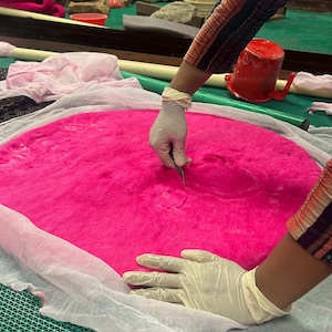 Peut inclure: Un grand tapis circulaire en laine feutr&eacute;e rose vif pos&eacute; sur un tissu en maille blanc. Un outil est utilis&eacute; pour presser le centre de la mati&egrave;re rose. La surface est textur&eacute;e et dense, pr&eacute;sentant une couleur satur&eacute;e et uniforme.