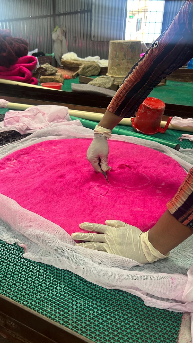 Peut inclure: Un grand tapis circulaire en laine feutr&eacute;e rose vif pos&eacute; sur un tissu en maille blanc. Un outil est utilis&eacute; pour presser le centre de la mati&egrave;re rose. La surface est textur&eacute;e et dense, pr&eacute;sentant une couleur satur&eacute;e et uniforme.