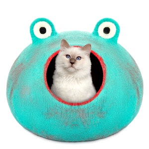 May include: A turquoise blue cat bed shaped like a frog with a white cat inside. The cat bed has red trim around the opening and black eyes on the frog's face.