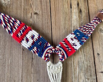 May include: A woven horse breast collar featuring a V-shaped design with red, white, and blue braided rope patterns. The two upper sections connect to a central metal ring, which attaches to a solid cream-coloured woven rope drop piece at the bottom.