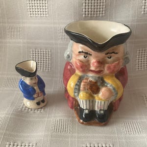 May include: Two ceramic figurines depicting a man in historical attire. The larger figurine is a pitcher with a black hat, red coat, yellow vest, and striped pants. The smaller figurine is a miniature version in blue and white.