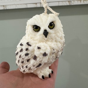 May include: White owl candle with black markings, detailed feathers, and yellow eyes. The candle is on a hand and has a white string attached to the top.