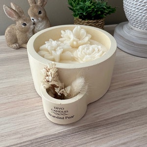 Puede incluir: Un portavelas de hormigón blanco con forma de taza con asa. La vela está perfumada con pera al brandy y presenta tres flores de cera blanca. El portavelas tiene un pequeño compartimento lateral con flores secas.