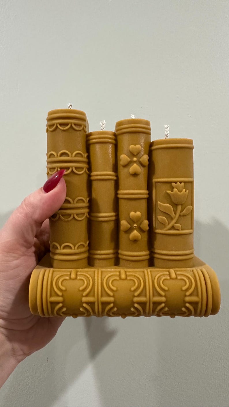 May include: A decorative candle set shaped like a stack of books. The candles are a warm, golden-brown color and feature detailed designs, including floral and geometric patterns. The base is also decorated with ornate details.