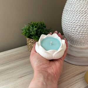 Op de afbeelding: Een witte keramische lotusbloemvormige kandelaar met een lichtblauwe kaars. De kaars heeft een enkele lont. De houder wordt in een hand gehouden.