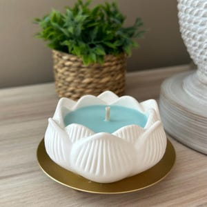 Op de afbeelding: Een witte lotusbloemvormige kandelaar met een lichtblauwe kaars erin. De kaars staat op een goudkleurige basis. Een kleine groene plant in een geweven mand staat op de achtergrond.