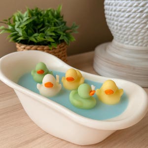 May include: A white ceramic miniature bathtub filled with light blue wax, featuring five small rubber duck figures in yellow, white, and light green colors with orange beaks. The ducks are arranged across the surface of the blue wax.