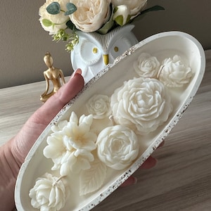Peut inclure: Plat ovale en céramique blanche avec un bord texturé, rempli de bougies blanches en forme de fleurs. Les bougies sont disposées en forme de bouquet.