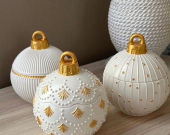 Puede incluir: Tres recipientes decorativos esféricos blancos con detalles dorados. Los recipientes tienen superficies texturizadas con varios patrones, incluyendo líneas verticales, puntos y diseños en forma de hojas. Cada recipiente tiene un lazo dorado en la parte superior.