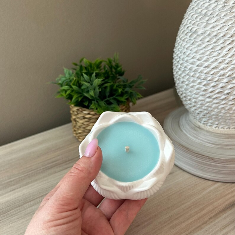 Op de afbeelding: Een witte, schelpvormige kandelaar met een lichtblauwe kaars. De kaars heeft een enkele lont. De houder wordt vastgehouden in een hand met roze nagellak. Een kleine plant in pot en een decoratieve lamp staan op de achtergrond.