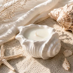 May include: A white ceramic candle holder shaped like a conch shell, filled with white wax and a single centered wick. The shell has a glossy finish and detailed ridges, positioned alongside a starfish and various natural seashells.