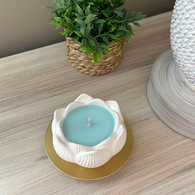 Op de afbeelding: Een witte keramische lotusbloem kaarsenhouder met een lichtblauwe kaars. De kaars staat op een goudkleurige basis. Een kleine groene plant in een geweven mand staat op de achtergrond.